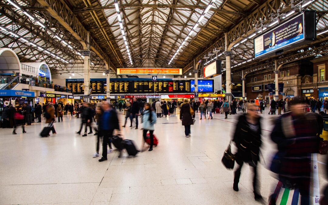 Modern railway station concourse: A bustling train station with a high metal roof, bright overhead lighting, electronic information boards, and many travelers walking through the busy space with luggage and shopping bags.